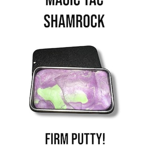 Könnte beinhalten: Eine schwarze Metallbox mit lila und grüner Knete. Der Text "MAGIC TAC SHAMROCK" und "FIRM PUTTY!" ist in Schwarz auf weißem Hintergrund gedruckt. Die Knete hat ein marmoriertes Aussehen.