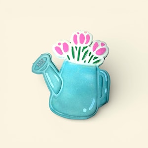 May include: A teal watering can-shaped decoration with a bouquet of pink and white flowers. The watering can has a handle and a spout with small holes. The flowers have green stems and are in full bloom.