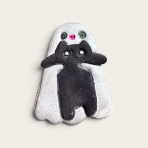 May include: A flat, sculpted ornament featuring a shimmering, pearlescent white ghost figure with two dark eyes and a small pink heart-shaped mouth. A solid black cat figure with two white dot eyes is layered over the center of the ghost.