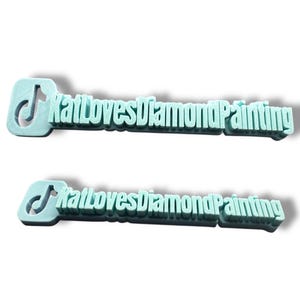 Puede incluir: Dos logotipos impresos en 3D de color azul claro. Cada logotipo presenta un icono de nota musical y el texto "KatLovesDiamondPainting". Los logotipos están ligeramente elevados, proyectando una sombra.