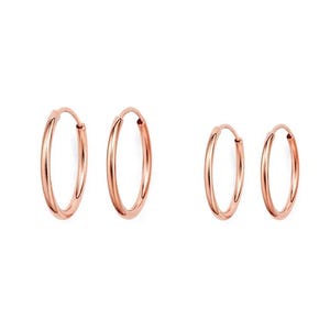 May include: Four rose gold hoop earrings of varying sizes are displayed against a white background. The earrings are simple, circular designs, reflecting light. The hoops are a warm, metallic color, suggesting a jewelry product.