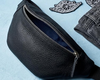 May include: A black pebbled leather fanny pack with a silver metal zipper closure and a dark blue interior lining. The bag features an adjustable black strap and is positioned next to a pair of dark gray denim jeans and two Jordan logo patches.