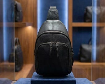 Pode incluir: Uma mochila transversal de couro preto granulado com formato de gota, um compartimento principal com zíper vertical e um bolso frontal menor com zíper. A bolsa possui ferragens de metal preto correspondentes e uma única alça superior.