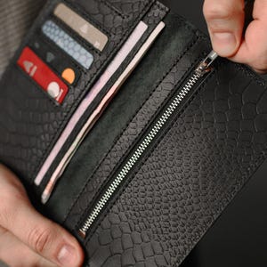 Puede incluir: Cartera de cuero negro con un patrón texturizado similar al de un reptil. La cartera está abierta, revelando múltiples ranuras para tarjetas llenas de tarjetas y un compartimento con billetes doblados. Una cremallera plateada recorre el borde.