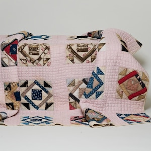 May include: A patchwork quilt featuring a light pink grid-stitched background with various square blocks. Each block contains geometric patterns in shades of blue, brown, red, and cream, showcasing diverse fabric prints and textures throughout the design.