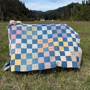 May include: A patchwork quilt featuring a grid of square fabric blocks in various colors including blue, yellow, pink, and white. The quilt displays a mix of solid colors and small-scale patterns, including stripes, checks, and floral prints.