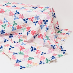May include: A white quilt with a star pattern in shades of red, blue, pink, and green. The quilt is draped over a surface, showcasing its intricate design and scalloped edges. The quilt's design features a repeating pattern of colorful stars.