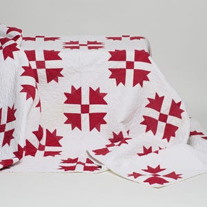 May include: A white quilt featuring a repeating pattern of deep red, star-shaped geometric motifs. The fabric has a visible quilted texture with intricate stitching throughout, creating a soft, layered appearance across the surface of the textile.