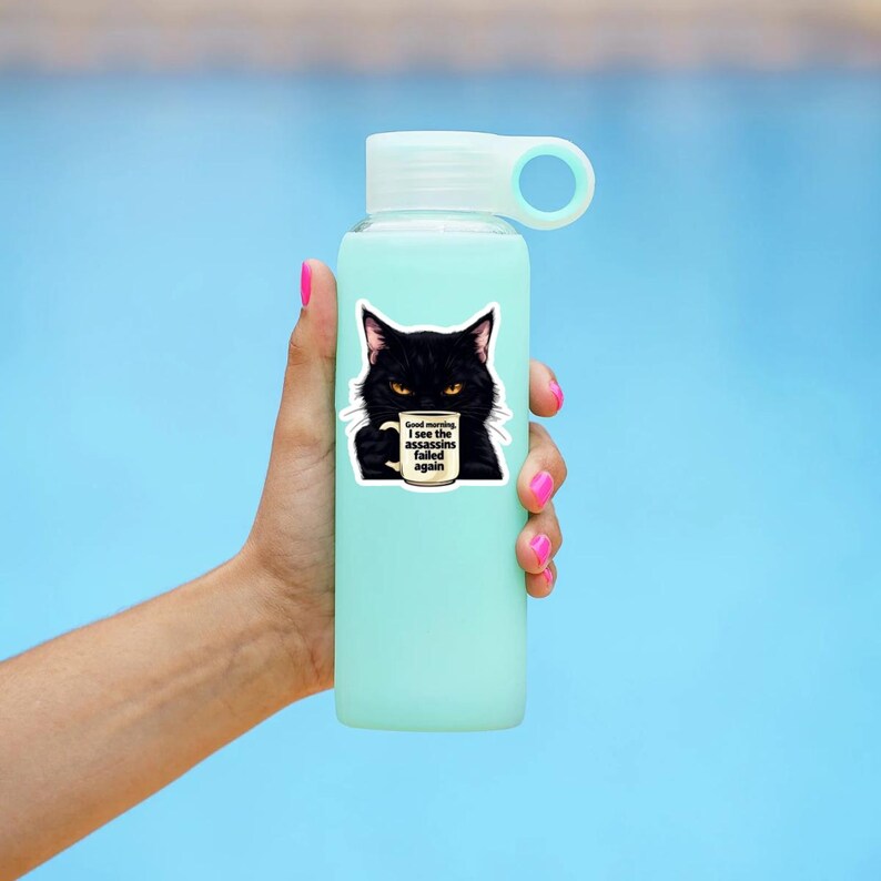 May include: A light blue water bottle with a white cap. A sticker of a black cat holding a coffee mug is on the bottle. The mug has the text "Good morning, I see the assassins failed again."