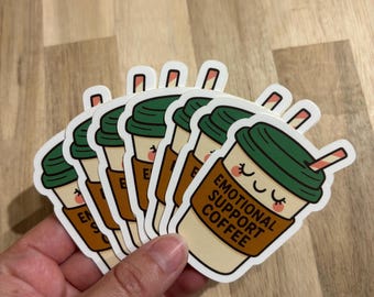 May include: A stack of stickers featuring a cartoon coffee cup with a green lid, brown sleeve, and a smiling face. The text on the cup reads "EMOTIONAL SUPPORT COFFEE". The stickers are held in a hand.