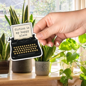 May include: A black typewriter sticker with gold keys and the text "fiction is my happy place" is held in front of several potted plants. The plants are on a wooden table near a window.