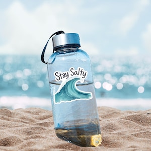 May include: A clear water bottle with a silver cap and a black strap. The bottle has a sticker that says "Stay Salty" with a blue wave design. The bottle is on a sandy beach with the ocean in the background.