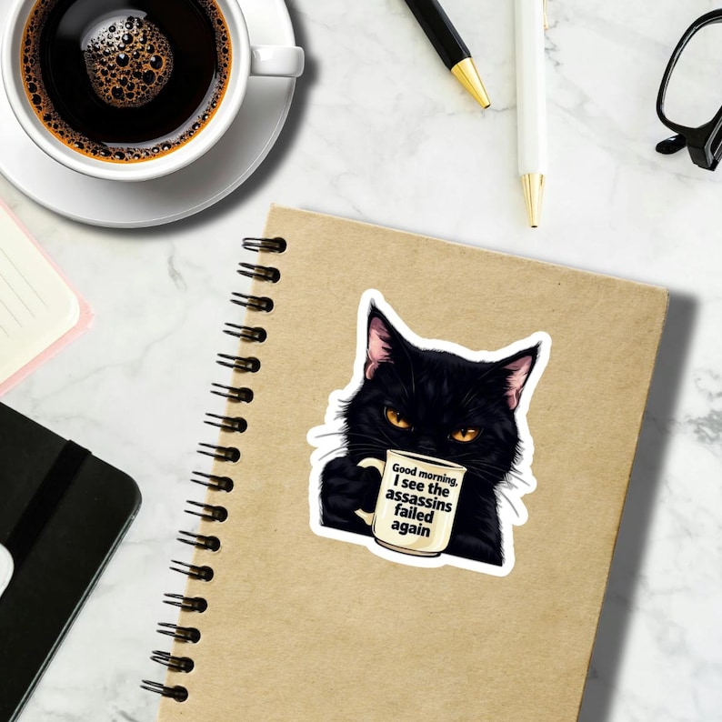 May include: A sticker featuring a black cat with yellow eyes holding a coffee mug. The mug has the text "Good morning, I see the assassins failed again." The sticker is on a brown notebook.