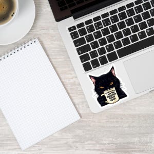 May include: A black cat sticker on a laptop, holding a coffee mug with the text "Good morning I see the assassins failed again." A spiral-bound notepad and a cup of coffee are also visible.