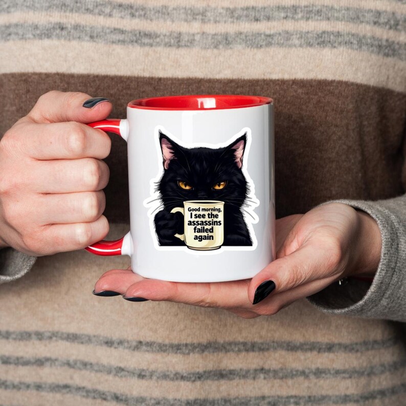 May include: White ceramic mug with a red interior and handle. The mug features a black cat illustration holding a coffee cup. The coffee cup has the text "Good morning, I see the assassins failed again."