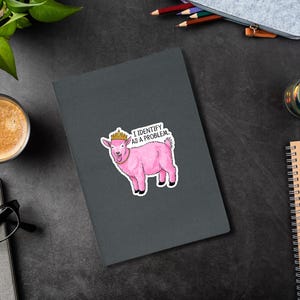 May include: A pink goat sticker with a gold crown and the text "I IDENTIFY AS A PROBLEM" on a dark gray notebook. The sticker is on a black desk with a cup of coffee, glasses, and pencils.