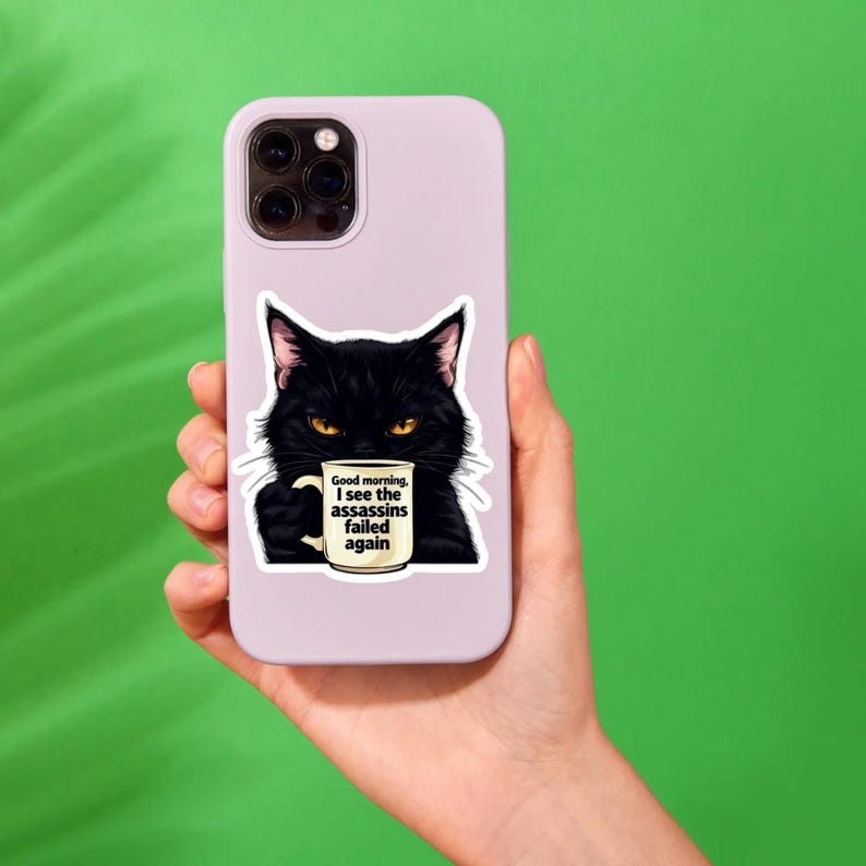 May include: A sticker featuring a black cat with yellow eyes holding a coffee mug. The mug has the text "Good morning, I see the assassins failed again." The sticker is on a lavender phone case, held against a green background.