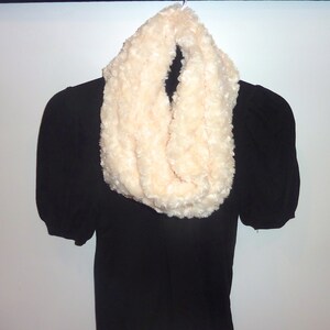 May include: A cream-colored, faux fur infinity scarf. The scarf is looped and has a soft, textured appearance. It is displayed on a black garment against a white background.