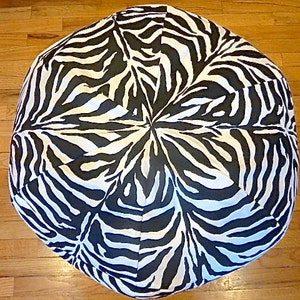May include: Round ottoman with a black and white zebra print pattern. The ottoman is a neutral color and would fit in any room. The zebra print is a classic design that adds a touch of style to any space.