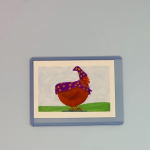 May include: A rectangular art print encased in a clear plastic sleeve featuring a painting of a brown chicken wearing a purple cape and a matching pointed hat, both decorated with yellow star patterns, set against a light blue and green background.