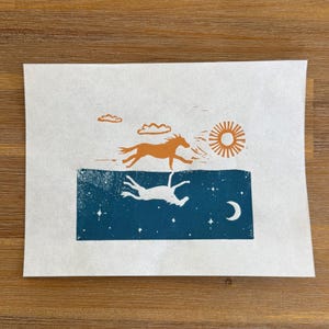 May include: A print depicting a horse running under a sun and clouds, with a mirrored image of the horse below in a night sky with stars and a crescent moon. The artwork is in shades of orange, white, and blue.