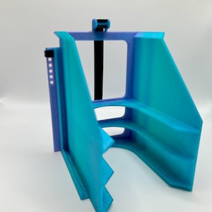 May include: A 3D-printed object in shades of blue and teal, featuring a rectangular frame with an open center and angled sides. A black strap and small black clip are visible. The object appears to be a support or holder.