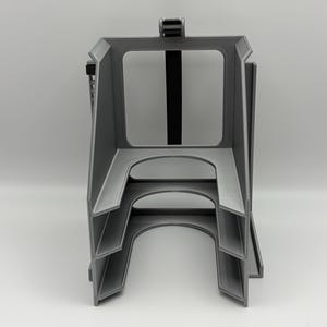 May include: A gray 3D-printed object with a rectangular frame and multiple levels. A black strap is attached to the top, and the object has a series of curved and angular cutouts. The design appears to be a holder or organizer.
