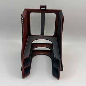 May include: A 3D-printed, geometric object with a red and black color scheme. The structure has an open rectangular top, a central vertical support, and arched lower sections. The design features layered edges and a textured surface.