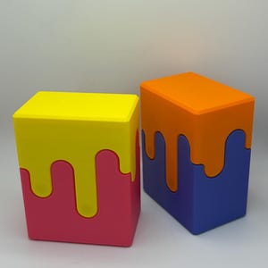 May include: Two colorful, cube-shaped boxes. One has a yellow top with pink sides and dripping yellow accents. The other has an orange top with blue sides and dripping orange accents. The boxes are side by side.