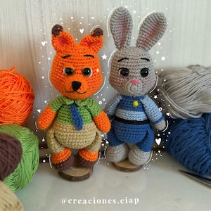 Puede incluir: Dos muñecos de personajes de ganchillo, un zorro y un conejo, están uno al lado del otro sobre bases de madera. El zorro es naranja con una camisa verde y pantalones color canela. El conejo es gris con un atuendo azul. Hilo y ganchillos en el fondo.