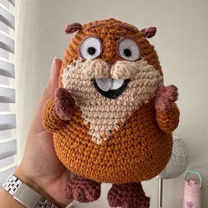 Pode incluir: Um brinquedo de pelúcia feito à mão, de crochê, em forma de personagem de desenho animado. O brinquedo apresenta um corpo marrom, rosto bege e detalhes rosa nas orelhas, mãos e pés. Possui olhos brancos grandes, boca sorrindo e expressão amigável.