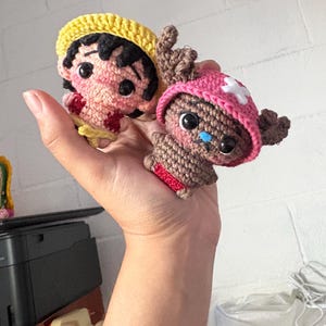 Puede incluir: Dos muñecos de personajes de anime hechos a ganchillo. Uno tiene un sombrero amarillo y pelo negro, el otro un sombrero rosa con una cruz blanca. Ambos muñecos tienen ojos negros y están hechos de hilo marrón, rosa y rojo.