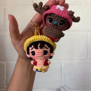 Puede incluir: Dos llaveros de ganchillo con personajes de anime. Uno tiene un sombrero amarillo, pelo negro, camisa roja y pantalones cortos azules. El otro es marrón con un sombrero rosa y cuernos. Hechos a mano y detallados.