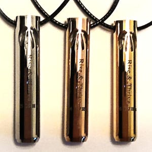 May include: Three metallic pendant necklaces in silver, bronze, and gold, each suspended from a black cord. The pendants are cylindrical with a carved design and the words "Rise & Thrive" and "7B" engraved on them. The background is a light cream colour.