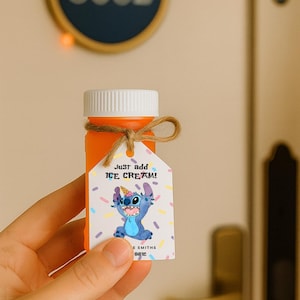 Può includere: Una bottiglia arancione con tappo bianco ed etichetta legata con spago. L'etichetta presenta un personaggio dei cartoni animati blu e la scritta "Just add ICE CREAM!". Lo sfondo include una porta con il numero 8602 e una stringa di luci.