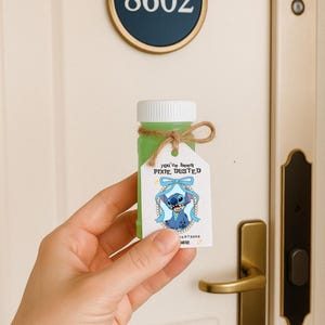 May include: A small, clear bottle filled with green liquid, with a white cap and twine tie. A tag with a cartoon character and the text "you've been PIXIE DUSTED" is attached. The bottle is held in front of a door with the number 8602.