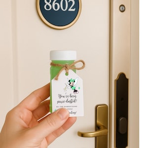 Op de afbeelding: Een kleine fles gevuld met groene vloeistof, een witte dop en een label vastgebonden met touw. Het label heeft een Minnie Mouse-ontwerp en de tekst "You've been pixie dusted!" Een deur met nummer 8602 op de achtergrond.