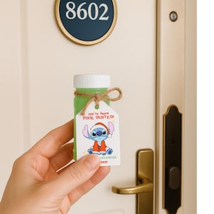 以下が含まれることがあります： 白いキャップが付いた小さな緑色のボトルと、漫画のキャラクターが描かれたタグ。タグには「Have a Roaring Good Time!」と書かれており、サンタの帽子をかぶった青いエイリアンが描かれています。ボトルは紐で結ばれています。