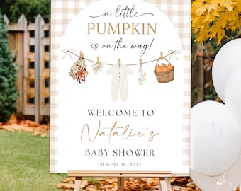 May include: A baby shower welcome sign on a wooden easel. The sign has a tan and white checkered background with the text "a little PUMPKIN is on the way!" and "WELCOME TO Natalie's BABY SHOWER AUGUST 16, 2025". The sign is decorated with illustrations of baby clothes and pumpkins.