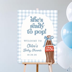 Peut inclure: Panneau de bienvenue pour une baby shower sur un chevalet. Le panneau a un fond vichy bleu et présente le texte "She's ready to pop!" et "Welcome to Chloe's Baby Shower 10.20.26". Une bouteille de cola de dessin animé est également présentée.