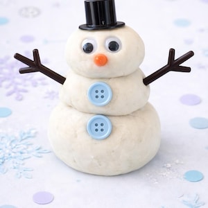 May include: A snowman made of dough, featuring a black top hat, googly eyes, and an orange nose. It has brown twig arms and light blue buttons. The snowman is a festive decoration.