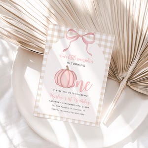 May include: A light pink and beige first birthday invitation with a pumpkin graphic. The text reads "Our little pumpkin is turning one." The invitation is on a white plate with a beige palm leaf in the background.
