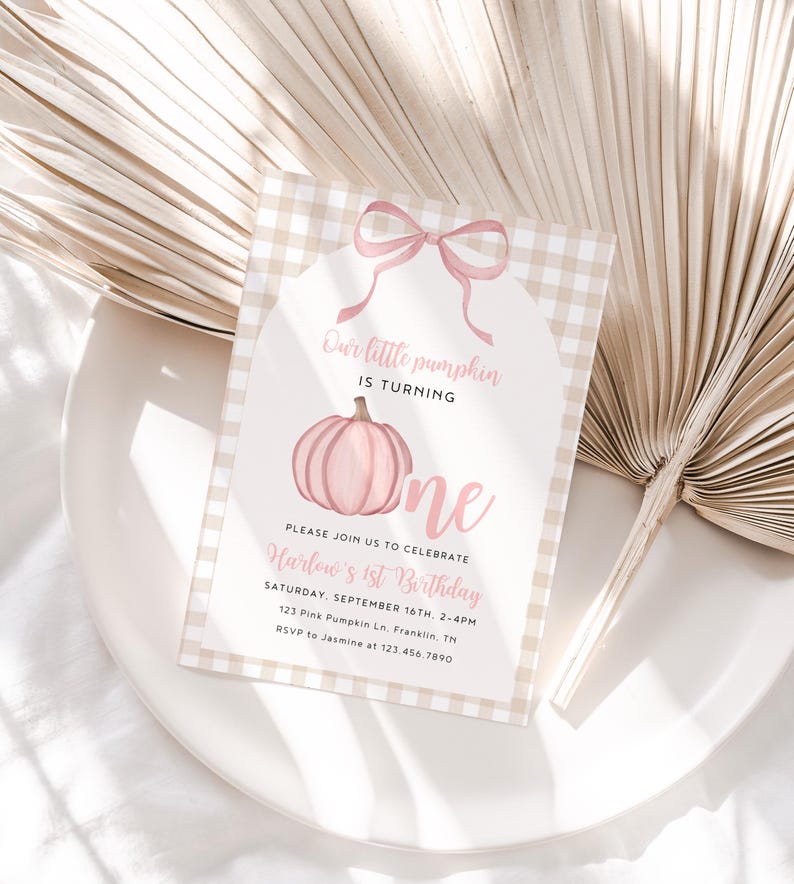 May include: A light pink and beige first birthday invitation with a pumpkin graphic. The text reads "Our little pumpkin is turning one." The invitation is on a white plate with a beige palm leaf in the background.