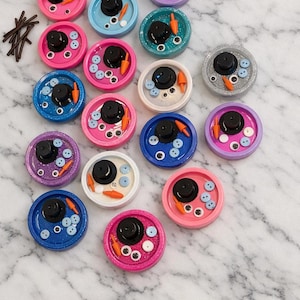 Pu&ograve; includere: Una collezione di colorati pezzi d'arte in resina a tema pupazzo di neve. Ogni pezzo presenta un cappello a cilindro nero, un naso di carota, occhi a bottone e dettagli di bottoni, su una base circolare glitterata.