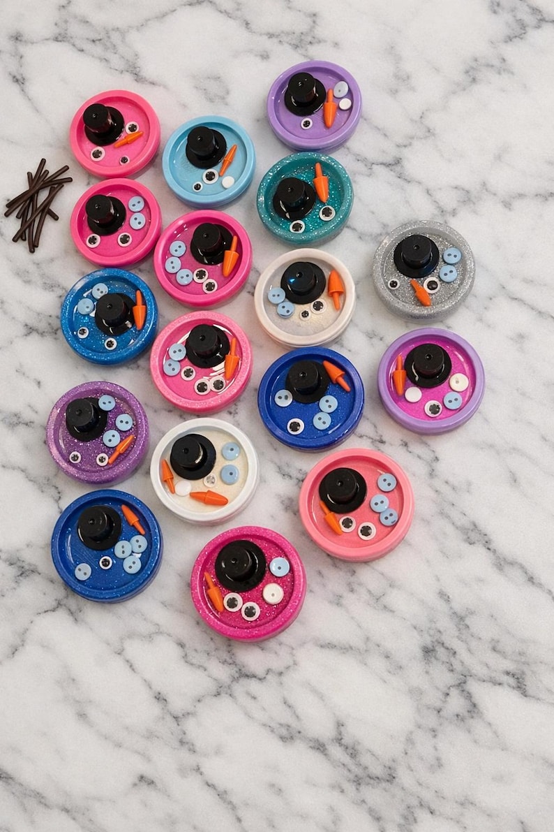 Pu&ograve; includere: Una collezione di colorati pezzi d'arte in resina a tema pupazzo di neve. Ogni pezzo presenta un cappello a cilindro nero, un naso di carota, occhi a bottone e dettagli di bottoni, su una base circolare glitterata.