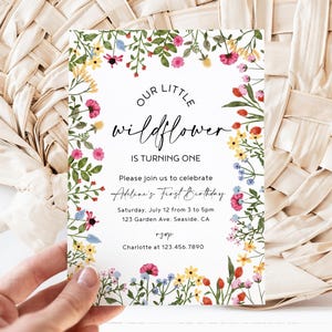 May include: A birthday invitation featuring the text "OUR LITTLE WILDFLOWER IS TURNING ONE." The invitation is decorated with a floral border of colorful flowers and foliage. The invitation includes the name "Addiline's First Birthday" and event details.