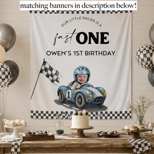 May include: A white fabric banner featuring a black checkered border, the text "OUR LITTLE RACER IS A fast ONE" and "OWEN'S 1ST BIRTHDAY", and a watercolor illustration of a child in a blue race car with a checkered flag.