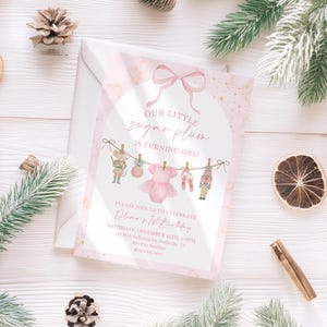 May include: A pink and white invitation card with the text "OUR LITTLE sugar plum IS TURNING ONE!" and details for a first birthday celebration. The card features illustrations of a mouse, a dress, ballet shoes, and a nutcracker.