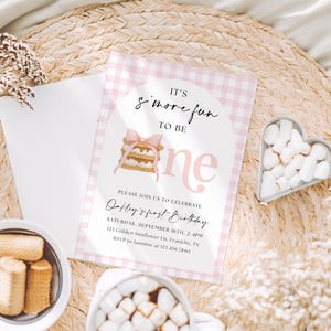Könnte beinhalten: Eine Geburtstagseinladung mit einem rosa Vichy-Karo-Rand und dem Text "It's s'more fun to be one". Die Einladung enthält eine Aquarellillustration eines S'more und eine rosa Schleife. Die Einladung ist von Marshmallows, Keksen und einer Tasse heißer Schokolade umgeben.