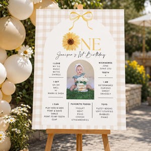May include: A rectangular birthday milestone sign with a beige gingham pattern, featuring a yellow sunflower, a gold bow, and the text "ONE" and "Juniper's 1st Birthday". It includes a central photo of a child and lists of personal facts.
